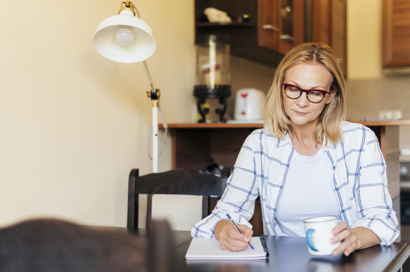 Mulher de uns 45 anos, de óculos, escrevendo num caderno em casa, com uma xícara de café ao lado