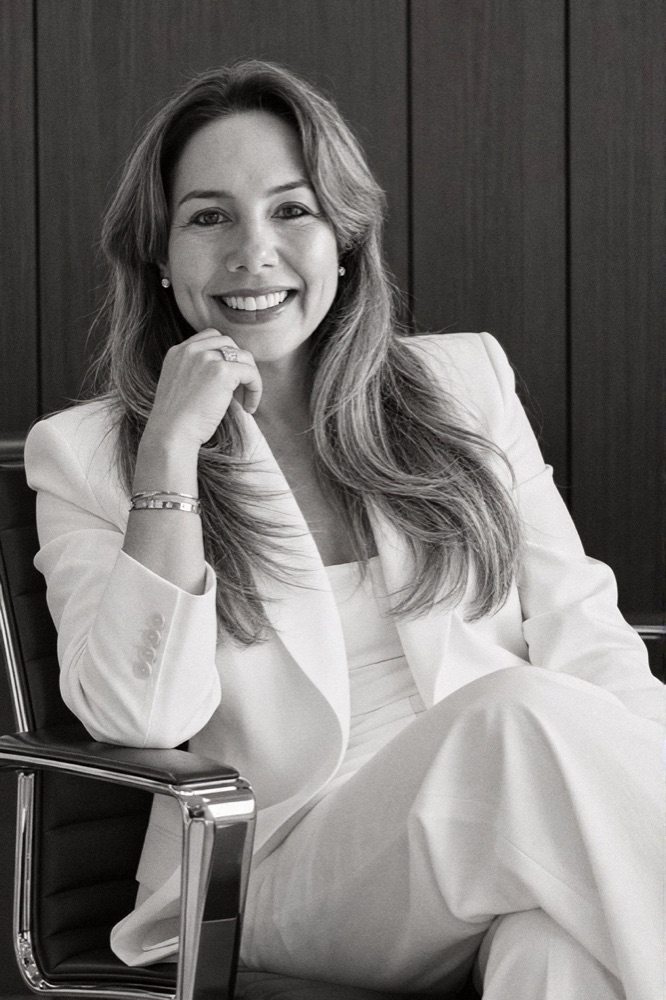 Retrato em preto e branco da Viviane Canziani, de terno claro, sentada em uma cadeira de escritório